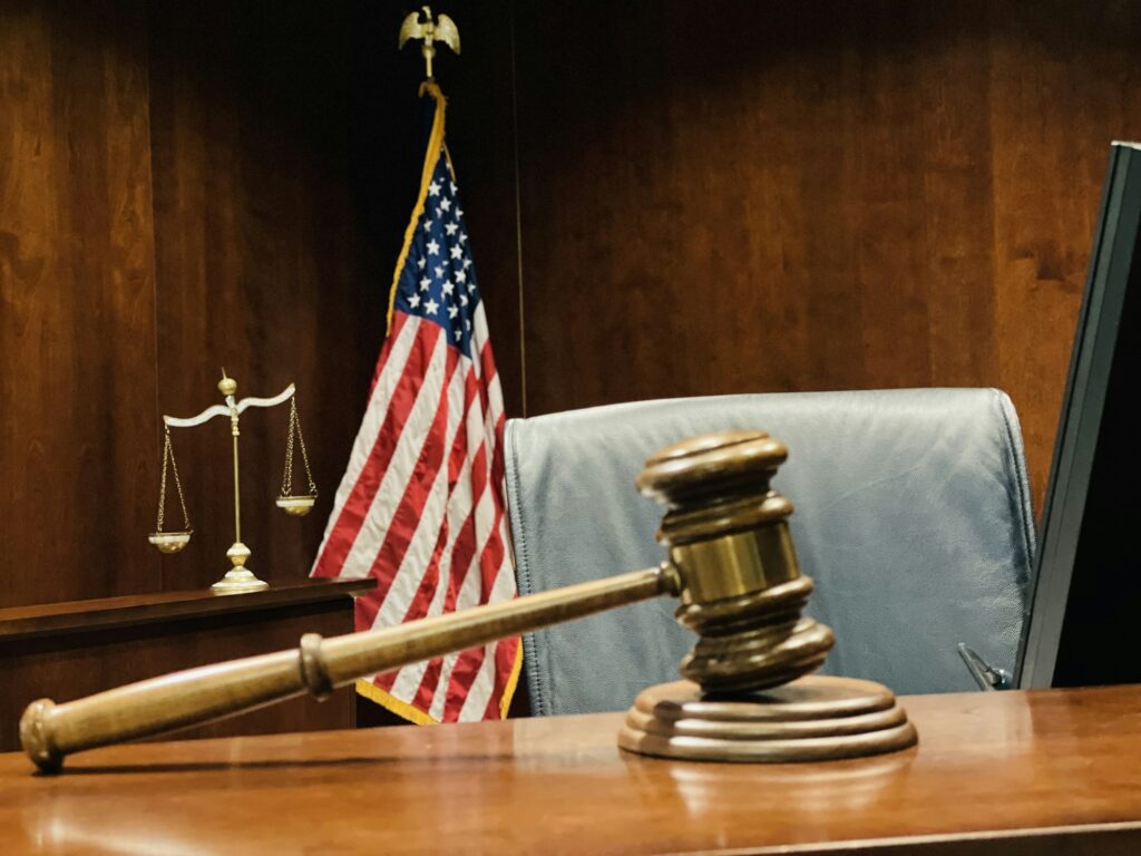 Judge’s wooden gavel on judicial bench, scales of justice and flag of US in background.
